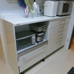 以前冷蔵庫と食器棚を置いてあったところに家電収納と４段引出し、ゴミ箱ストッカーもついてすっきりしました。吊戸とカウンターの間はキッチンパネルではなくマグネットボードです。廊下から見える壁には大胆なアクセントクロスで素敵なキッチンになりました。