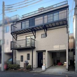 施工後の外観です。3階建ての建物は、ビルのようなシャープな形状を活かしつつ、外壁を明るめのグレーベージュで仕上げ、落ち着きのある印象に整えました。
2階の一部および3階バルコニーの手すりは黒色で塗装し、雨どいも同じく黒で統一。屋上まわりのスレート屋根部分には、軽量で耐久性に優れたスーパーガルテクトを使用し、カバー工法で施工しています。外壁とのコントラストが美しく、おしゃれなアクセントになっています。
また、屋上には防水塗装工事を施しており、建物全体の耐久性と美観を両立させました。