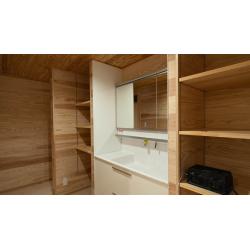 【木のぬくもりを感じる洗面・収納スペース】
全体を明るい木目の建材で統一し、自然で温かみのある空間に仕上げました。壁、天井、そして左右に配置された造作の収納棚に至るまで、美しい木目が連続することで、心地よく落ち着いた雰囲気を醸し出しています。
洗面化粧台は、清潔感のあるホワイトで木材とのコントラストをつけ、機能的な三面鏡収納を採用。洗面ボウル下にも引き出し収納を設け、散らかりがちな洗面周りのアイテムをすっきりと収納できます。

左右には、高さの調節が可能なオープンシェルフを造作。十分な収納量を確保しつつ、オープンにすることで圧迫感を軽減し、使い勝手の良い洗面・脱衣スペースを実現しました。

