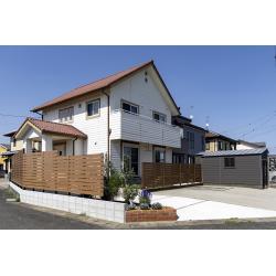 全体をブラウンでまとめ、可愛らしいお家の雰囲気に寄り添うナチュラル外構が完成しました。中古住宅での新生活に合わせて外回りを整えたいとのご相談から、既存コンクリート横に駐車場を新設し、横にはレンガ花壇で華やかさをプラス。奥にはインターロッキングを馬目地で敷いたアプローチを設け、玄関までの動線もやさしい印象に。横のお庭には人工芝が広がり、リビング前には出幅1.2ｍのデッキを設置。火を使うBBQのためにデッキ下は広めにコンクリート舗装とし、使い勝手と安全性を両立しています。お庭のフェンスも、デッキに腰掛けた際に視線が気にならない高さに整え、ほどよい囲われ感の落ち着く空間となりました。