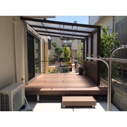 使用している商品はテラス屋根「ＳＭＡＬＥ」腰パネル付きです。柔らかい木目の色合いが緑との相性も良く、圧迫感無く目隠しをしてくれています。木樹脂デッキには、オプションでお子さんの転落防止のための柵をつけています。勝手口側にはコンクリートを打設し、これから雑草に悩まなくても大丈夫な素敵なお庭になりました。