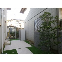 建物の雰囲気に合わせて木調のタイルを使用し、シンプルなテラスが完成です。隣地の窓を隠すためにブロックとフェンスを設置し、圧迫感がでないように樹木（ソヨゴ）を植えて空間を和らげています。まわりには人工芝を敷き、明るい雰囲気となりました。
