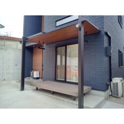 既存のウッドデッキの上にテラス屋根を施工。玄関の隣でしたので、ご希望は住宅にマッチする意匠性の高いテラス屋根。いくつかの候補から意匠性の高さ・屋根のスタイリッシュさ・施工方法・コストパフォーマンス等、様々な点からLIXILのテラスSC（木彫）を採用。住宅にポイントで入っている木調色とマッチしてとても自然でカッコいい仕上がりです。オプションで車庫空間を照らすセンサー照明とお洗濯に便利な物干しを採用しています。